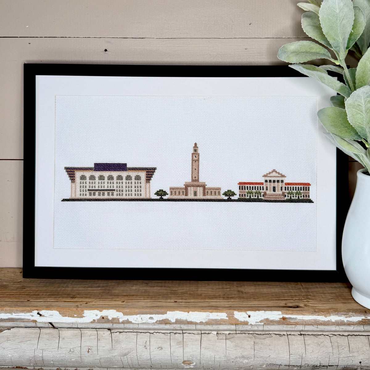 A framed cross stitch portrait of the officially licensed LSU cross stitch kit featuring a colorful architectural cityscape illustration, styled on a rustic wooden shelf next to a white ceramic vase with green leaves against a beige wood-paneled wall.